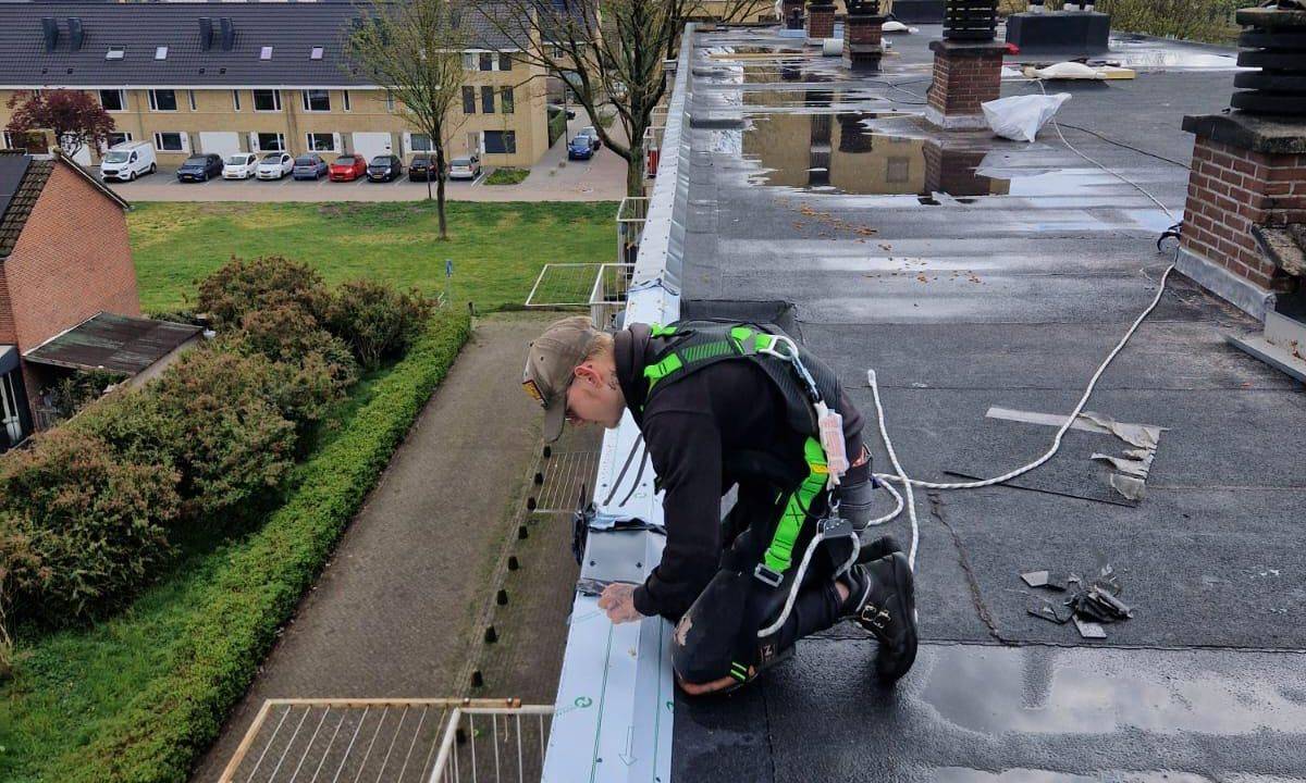 Dakdekker met veiligheidslijn werkt aan het plaatsen van dakrandafwerking op een plat dak van een appartementencomplex.