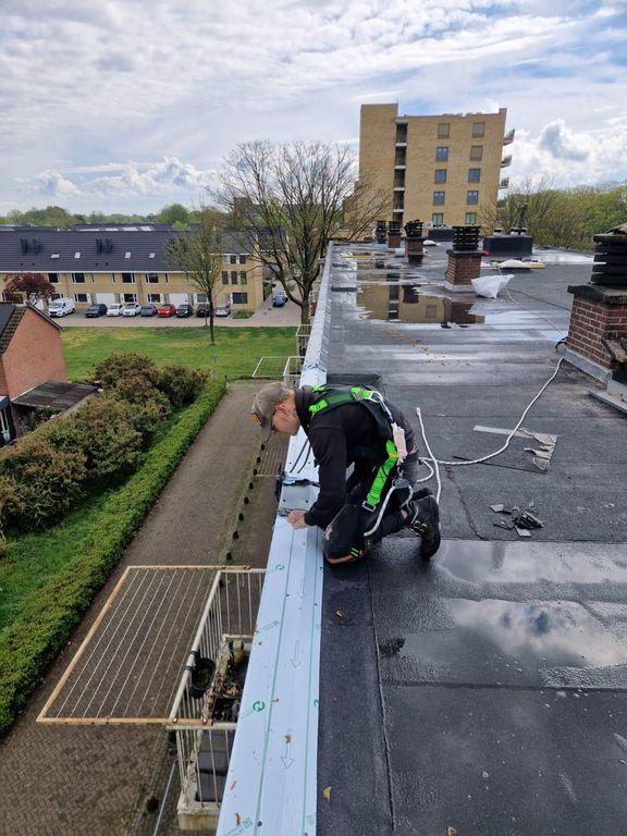 Dakdekker met veiligheidslijn werkt aan het plaatsen van dakrandafwerking op een plat dak van een appartementencomplex.