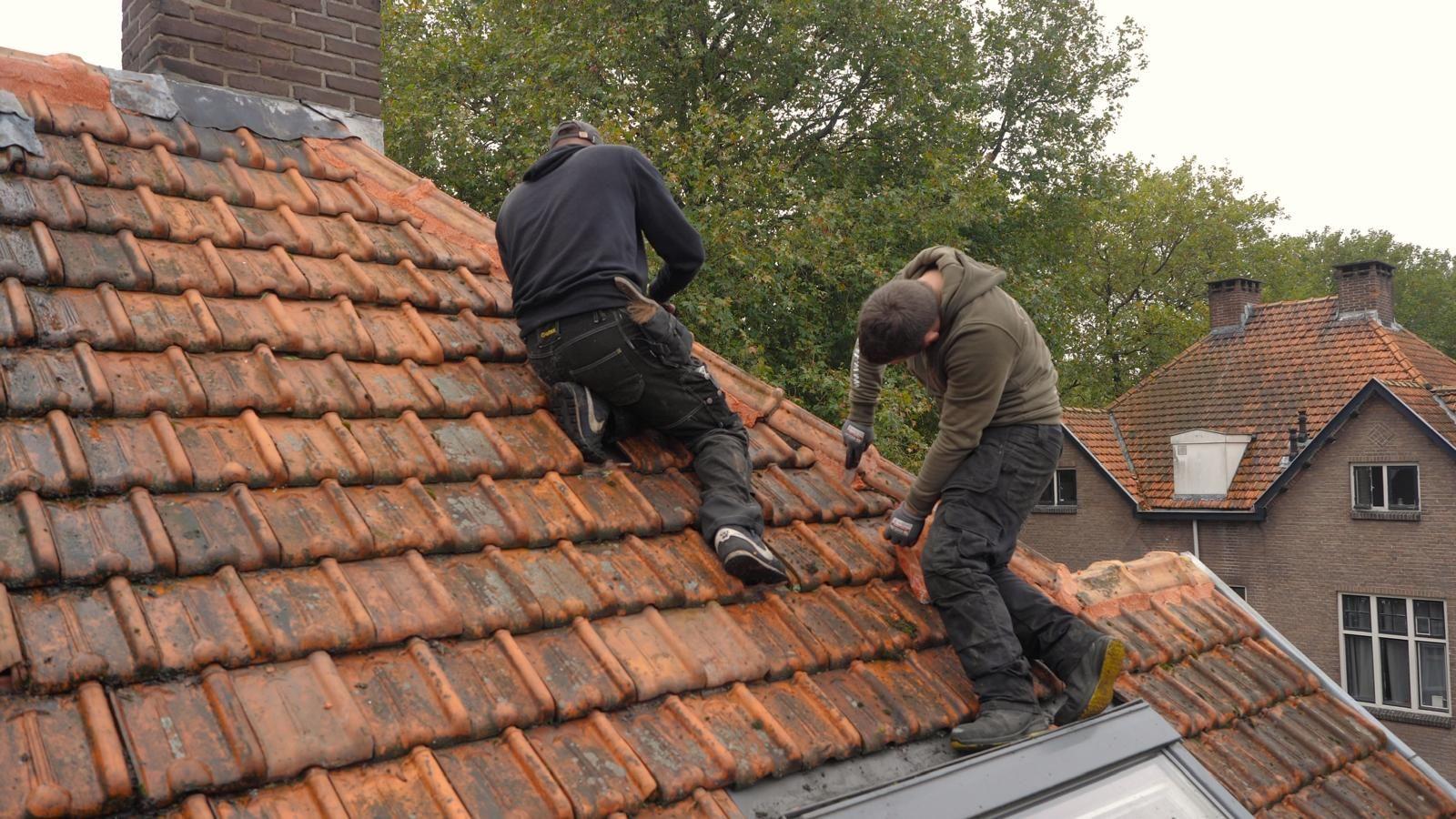 Twee dakdekkers zijn bezig met het herstellen van een oud pannendak, waarbij ze voegen en reparatiemateriaal aanbrengen terwijl ze gebukt op het schuine dak werken