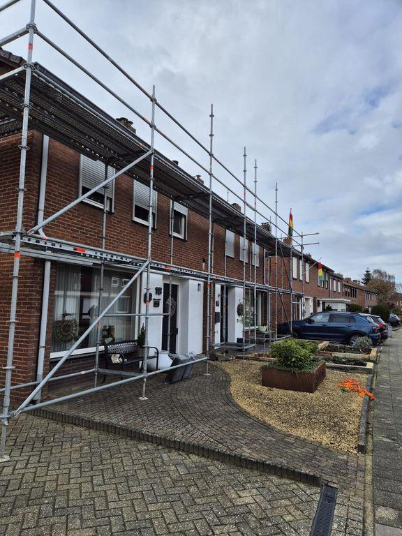 Een rij bakstenen woningen met witte gevelaccenten staat onder een bewolkte hemel volledig in de steigers voor renovatiewerkzaamheden.
