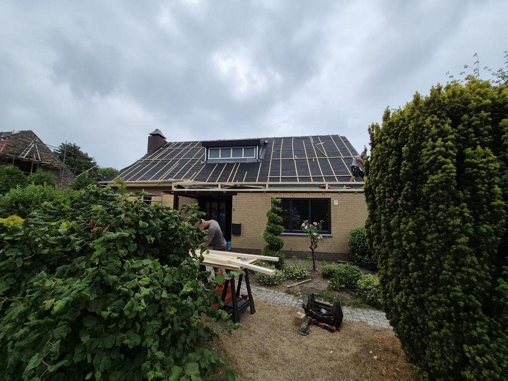 Een vrijstaande woning met een dakkapel toont het houten raamwerk van nieuwe panlatten op een donker onderdak, met op de voorgrond een aanhanger vol steigermateriaal.