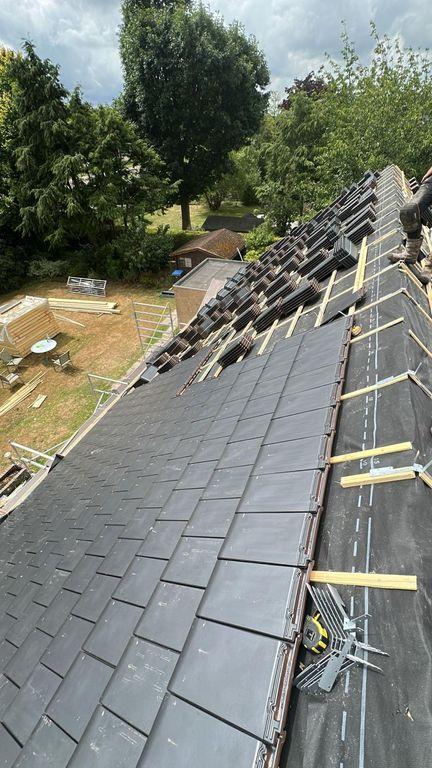  Een bovenaanzicht van een schuin dak in renovatie, waarbij een groot deel al is bedekt met moderne antracietgrijze dakpannen en stapels pannen klaarliggen op de panlatten voor verdere verwerking.