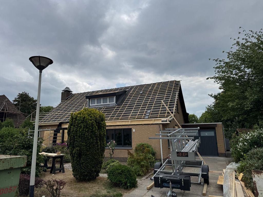  Een vrijstaande woning met een dakkapel toont het houten raamwerk van nieuwe panlatten op een donker onderdak, met op de voorgrond een aanhanger vol steigermateriaal.