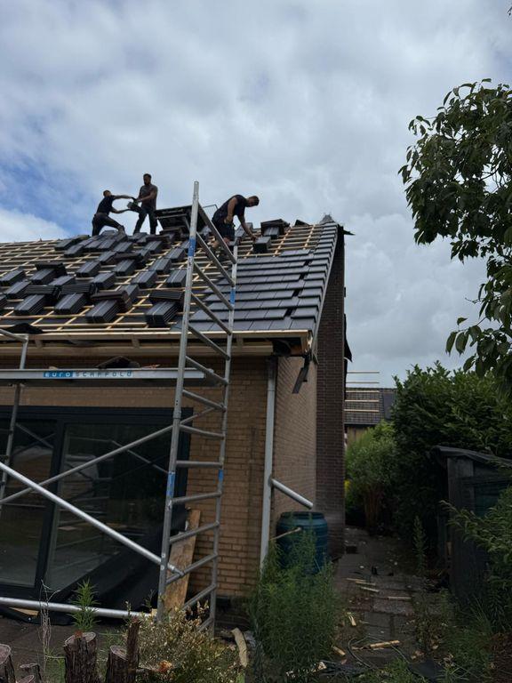 Drie dakdekkers zijn op een hellend dak aan het werk om nieuwe antracietgrijze dakpannen te leggen, terwijl een aluminium rolsteiger tegen de zijgevel van de woning staat.