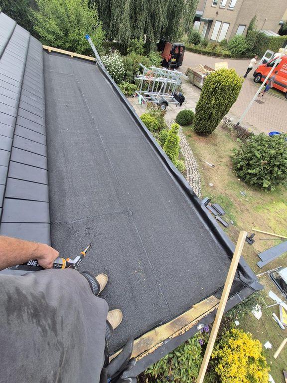 Een overzicht van een plat dak dat vers is voorzien van zwarte bitumineuze dakbedekking, met op de achtergrond een ladder en een werkbus.