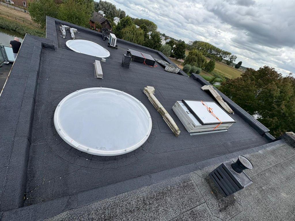  Een groot plat dak bedekt met zwart bitumen is voorzien van twee ronde lichtkoepels en diverse installaties, zoals zonnepanelen en een airconditioningunit.