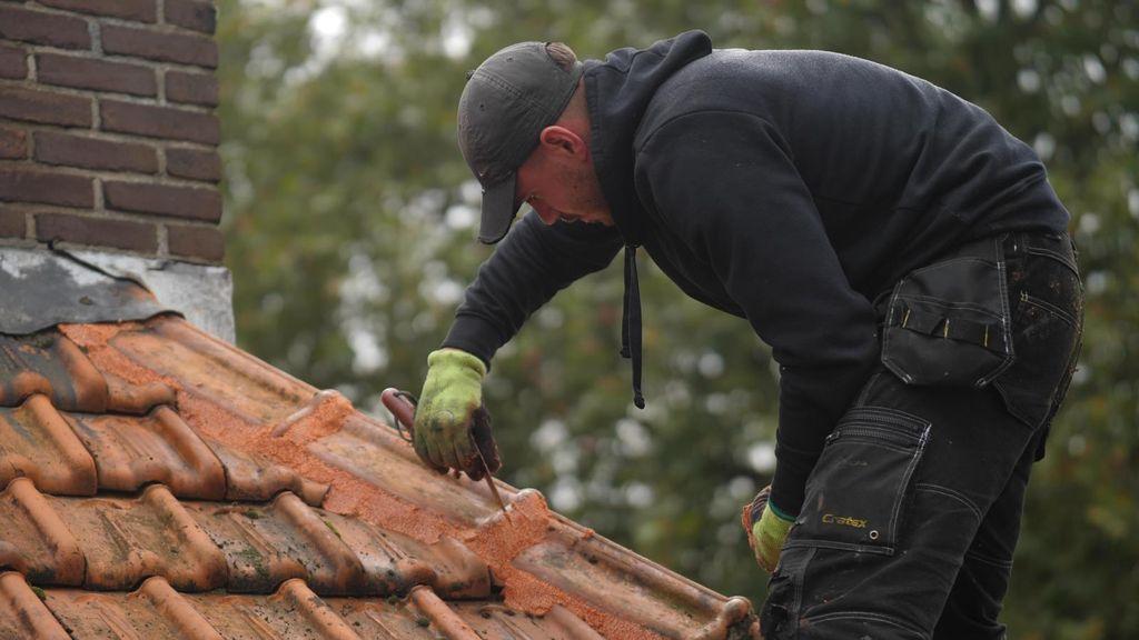 Dakdekker repareert een pannendak met voegmateriaal terwijl hij gebukt werkt bij een schoorsteen, met werkhandschoenen en gereedschap, in een buitenomgeving.