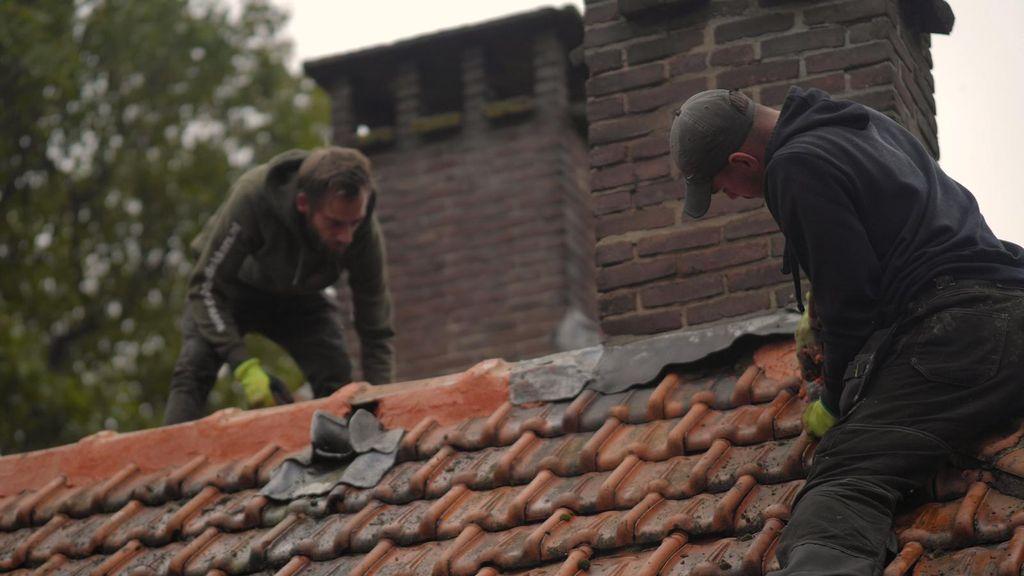 Twee dakdekkers herstellen een pannendak bij een schoorsteen, waarbij ze voegmateriaal en gereedschap gebruiken terwijl ze gebukt werken op het dak.